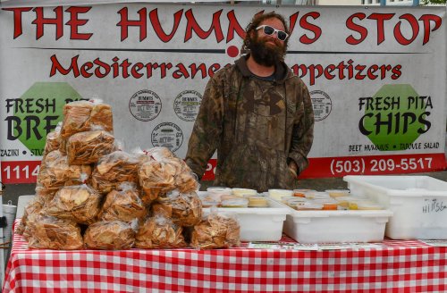 A bearded man in a camouflage hoodie stands behind a table with stacked pita chips and containers of hummus.