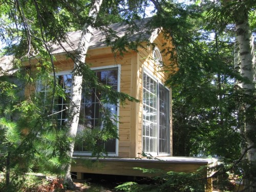 A small wooden cabin nestled among trees, featuring large windows and a sloped roof.
