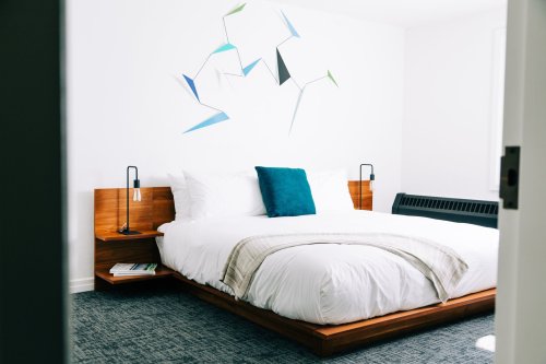 A modern bedroom featuring a wooden bed, white bedding, a blue accent pillow, and abstract wall art.