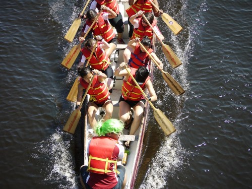 A team of rowers in a dragon boat, wearing life vests, paddling vigorously on a calm water surface.
