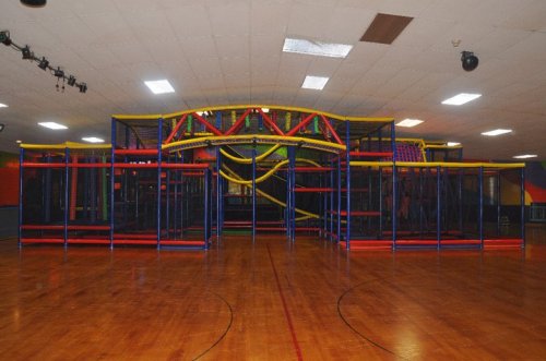Colorful indoor play structure with slides and climbing areas, set in a spacious gymnasium with wooden floors.