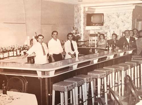 A vintage bar scene with bartenders and patrons, featuring a long counter and shelves of liquor bottles in the background.