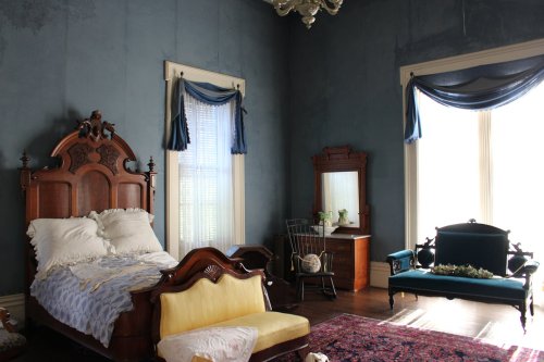 A vintage bedroom featuring a wooden bed, a yellow settee, a mirror, and blue walls with elegant drapes.