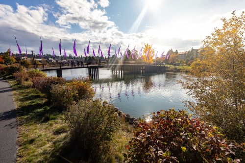 A scenic view of a lake with a wooden bridge and colorful flags, surrounded by trees and vibrant autumn foliage.