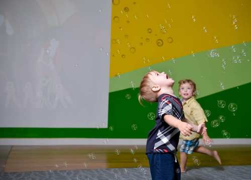Two young children play joyfully, reaching for floating bubbles in a colorful room with a projection in the background.