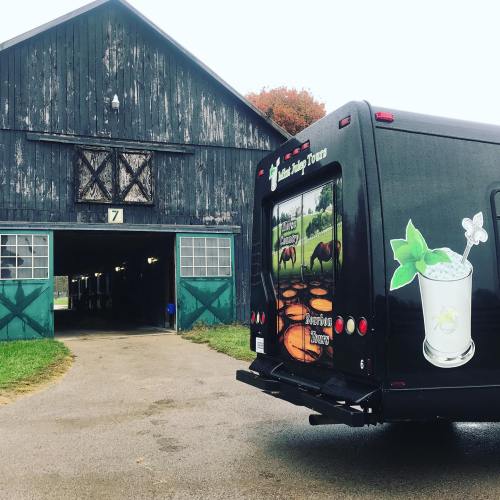 A black tour van parked near a rustic barn with green doors, surrounded by grass and trees in autumn colors.