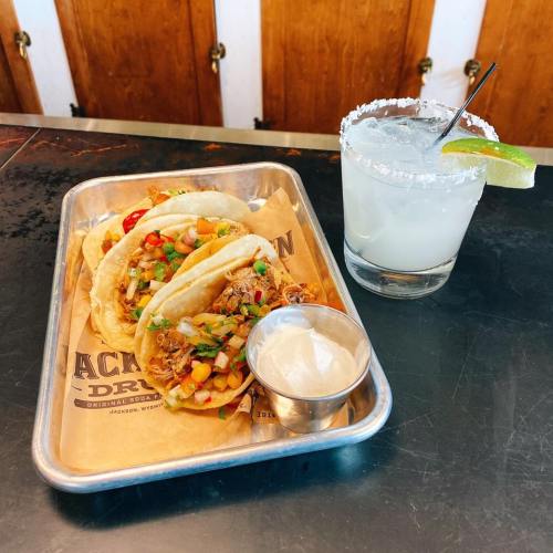 A metal tray with three tacos topped with vegetables, alongside a margarita with a lime wedge and salt rim.