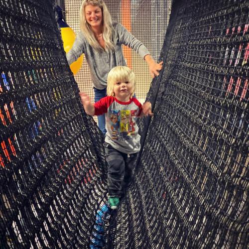 A child walks on a net bridge while an adult stands behind, both smiling in a playful indoor setting.