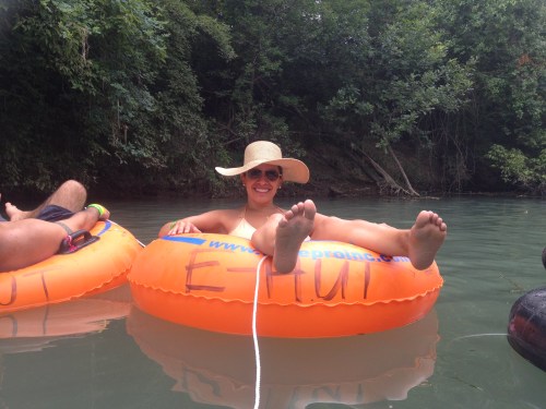 A person relaxes on an orange inner tube in a river, wearing a wide-brimmed hat and smiling, with feet visible.