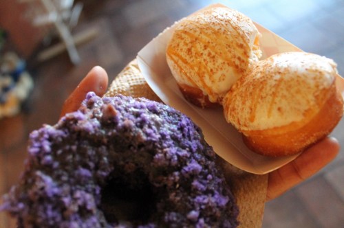 A hand holding a purple donut and two cream-filled pastries dusted with cinnamon sugar.