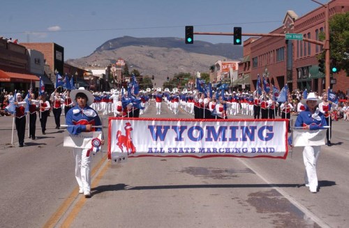 A marching band in white and blue uniforms parades down a street, holding a banner that reads "Wyoming All State Marching Band."