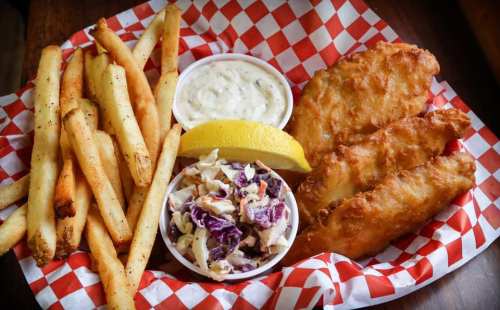 A basket of golden fried fish fillets, fries, coleslaw, and a lemon wedge, served with dipping sauce.