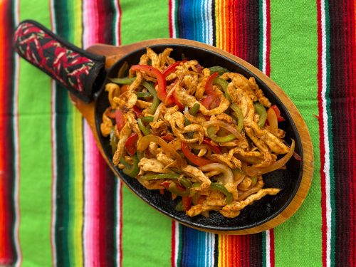 Sizzling chicken fajitas with bell peppers and onions on a wooden platter, set on a colorful striped tablecloth.