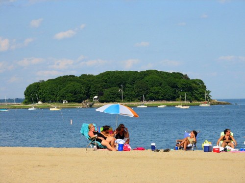 A sandy beach scene with people relaxing under umbrellas, with an island and boats in the background.