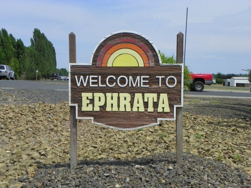 A wooden sign reading "Welcome to Ephrata" with a colorful sunrise design, surrounded by gravel and trees.