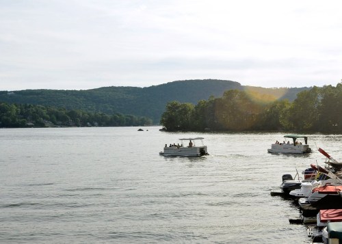 Two boats navigate a calm lake surrounded by green hills under a cloudy sky. Docked boats are visible in the foreground.