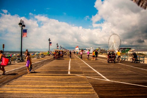 A bustling boardwalk with people walking, biking, and a ferris wheel in the background under a partly cloudy sky.