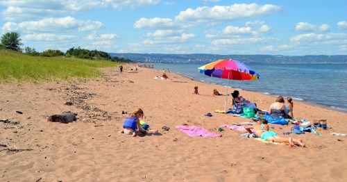 A sandy beach with people relaxing under colorful umbrellas, children playing, and a calm water view.