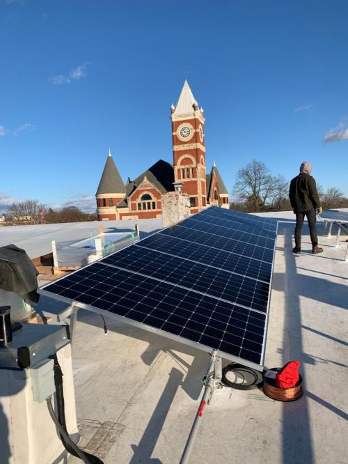 A person stands on a rooftop beside solar panels, with a historic clock tower in the background under a clear blue sky.