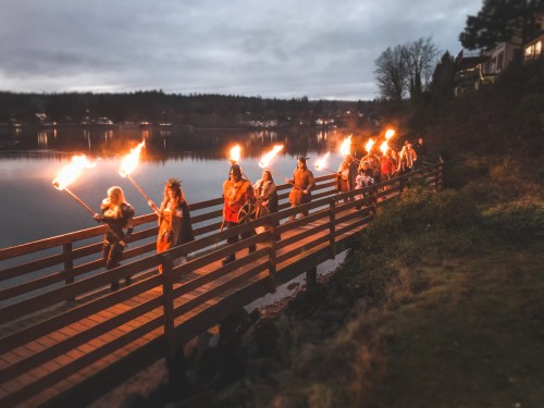 A group of people in costumes carrying torches walks along a wooden path by a lake at dusk.
