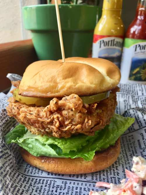 A crispy fried chicken sandwich with pickles and lettuce on a bun, served on a bed of newspaper with sauces in the background.