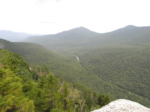 A panoramic view of lush green mountains and a winding road through a dense forest under a cloudy sky.