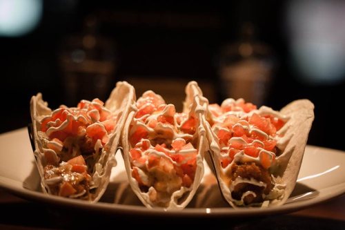 Three tacos filled with meat, topped with diced tomatoes and drizzled with creamy sauce, served on a white plate.