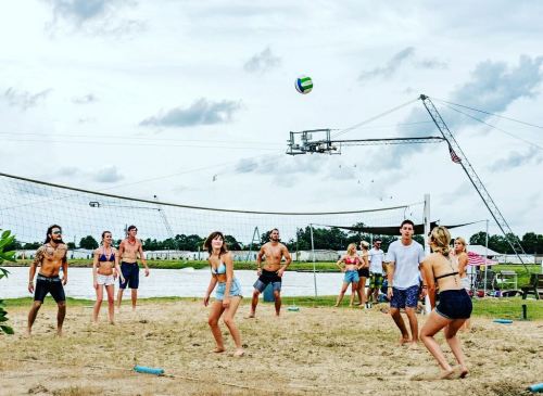 A group of people playing beach volleyball by a lake on a cloudy day, with water sports equipment in the background.