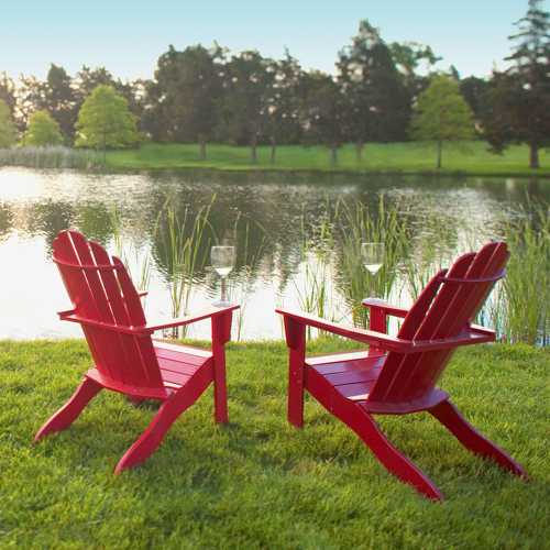 Two red Adirondack chairs facing a serene pond, surrounded by lush greenery and tall grasses.