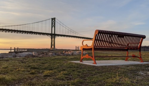 A red bench overlooks a river with a bridge in the background during sunset, surrounded by grass and rocks.