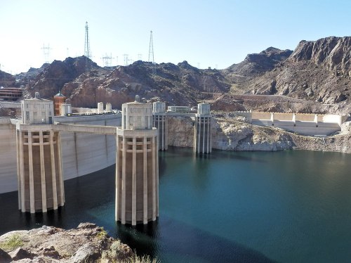 A large dam with two towers, surrounded by rocky terrain and a calm blue reservoir under a clear sky.