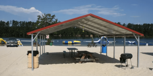 A beach shelter with a picnic table and grill, overlooking a lake with inflatable water toys and trees in the background.