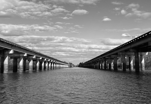 Black and white image of two parallel bridges over a calm river, with clouds reflecting in the water.