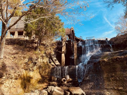 A watermill with a wooden wheel beside a cascading waterfall, surrounded by trees and a clear blue sky.