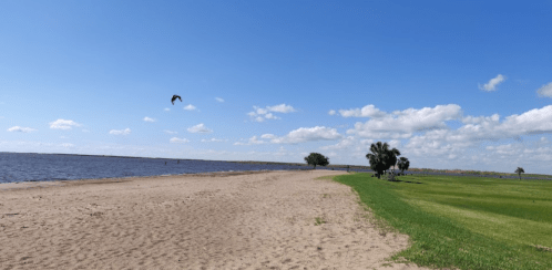 A sandy beach beside a calm lake, with green grass and a few trees under a blue sky with scattered clouds.