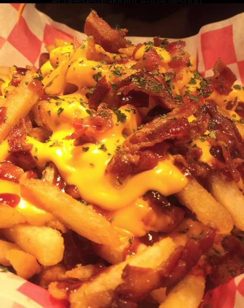 A close-up of loaded fries topped with melted cheese, crispy bacon bits, and a sprinkle of herbs in a red-checkered basket.
