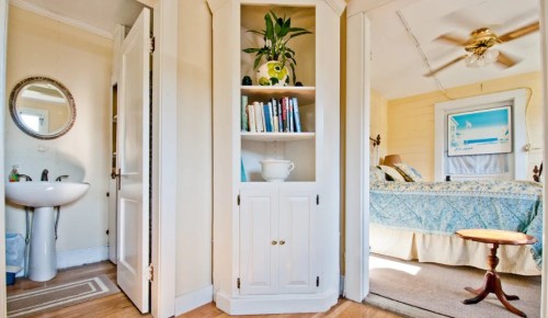A cozy room with a bookshelf, sink, and a view of a bedroom featuring a blue bedspread and a ceiling fan.