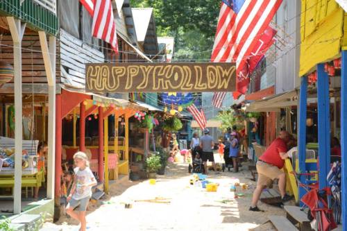 Colorful street scene in a festive village, featuring a "Happy Hollow" sign and American flags, with people enjoying the day.
