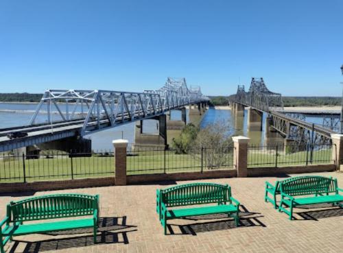 A view of two bridges spanning a river, with green benches in the foreground and clear blue skies above.