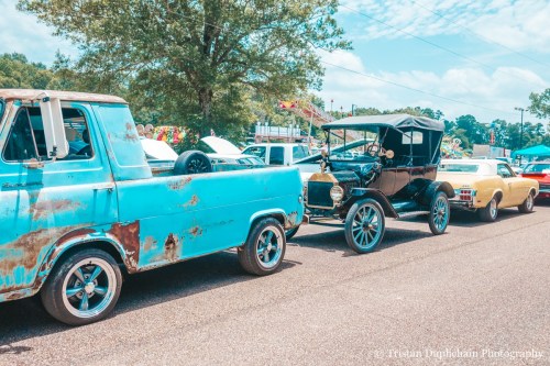 A row of vintage cars, including a blue truck and a classic black car, parked along a tree-lined street.