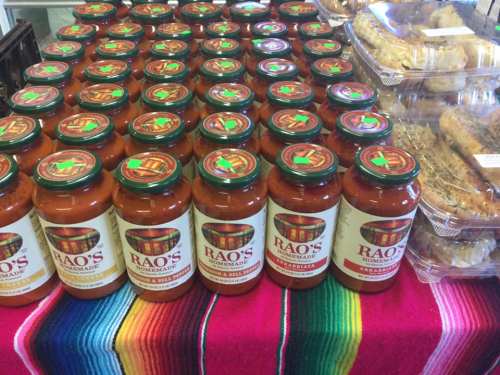 A display of Rao's marinara sauce jars arranged on a colorful tablecloth, with baked goods in the background.
