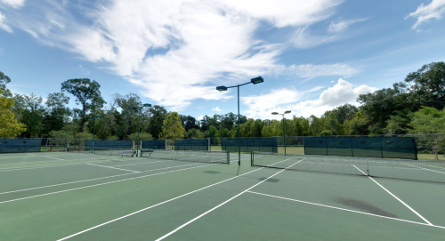 A sunny outdoor tennis court with green surfaces and surrounding trees under a blue sky with scattered clouds.
