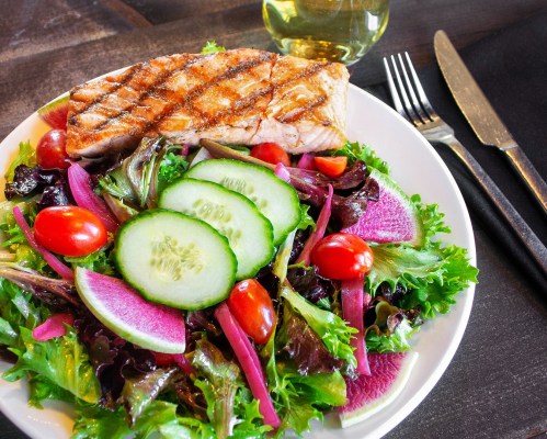 A plate of grilled salmon on a bed of mixed greens, cherry tomatoes, cucumber slices, and pink radish, with a glass of oil.