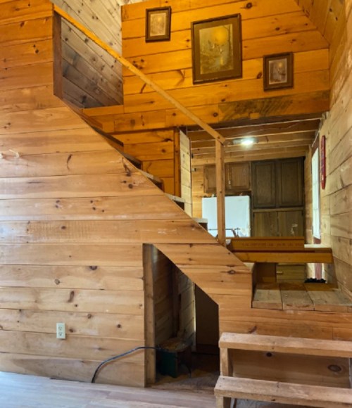 Interior of a wooden cabin featuring a staircase, wooden walls, and framed pictures. Warm, rustic atmosphere.