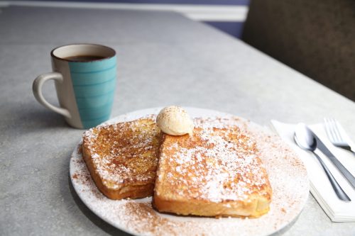 A plate of French toast topped with butter and powdered sugar, alongside a cup of coffee on a table.