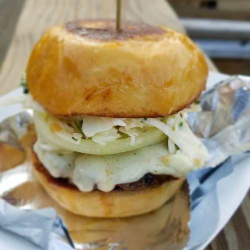 A close-up of a delicious burger with a shiny bun, layered with cheese, coleslaw, and pickles on a silver plate.