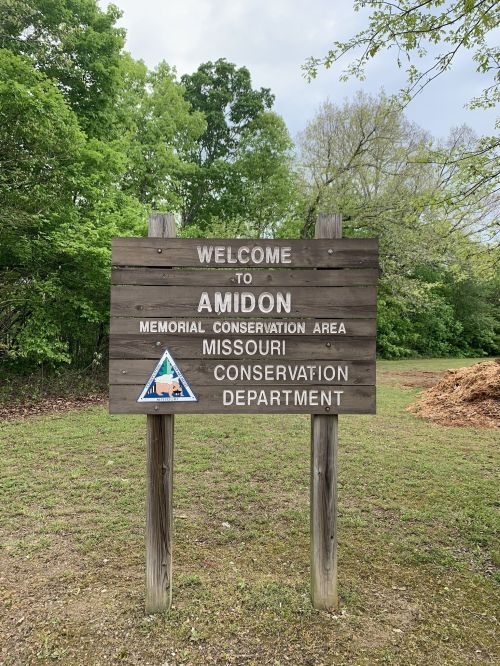Wooden sign welcoming visitors to Amidon Memorial Conservation Area, Missouri Conservation Department. Green trees in background.