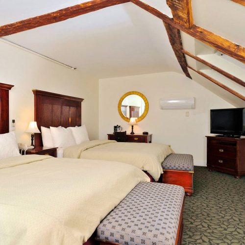 Cozy hotel room with two beds, a mirror, TV, and wooden beams on the ceiling. Warm, inviting decor.