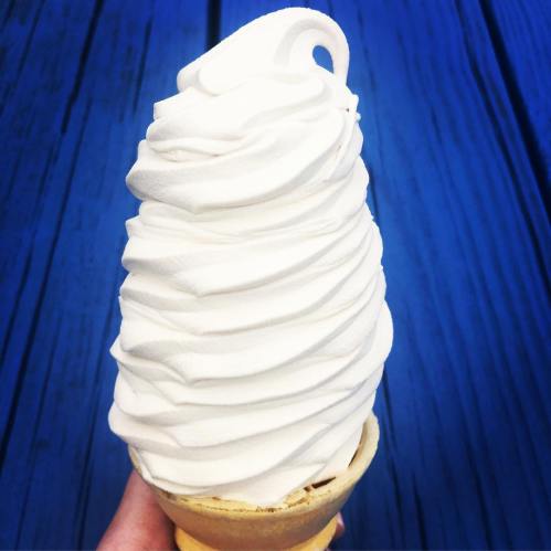 A hand holding a tall swirl of soft-serve ice cream in a cone, set against a blue wooden background.