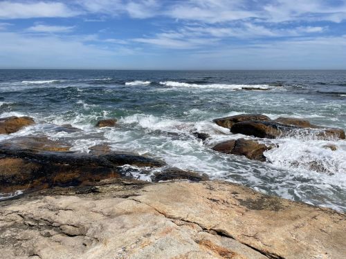 A rocky shoreline with waves crashing against the rocks under a blue sky with scattered clouds.